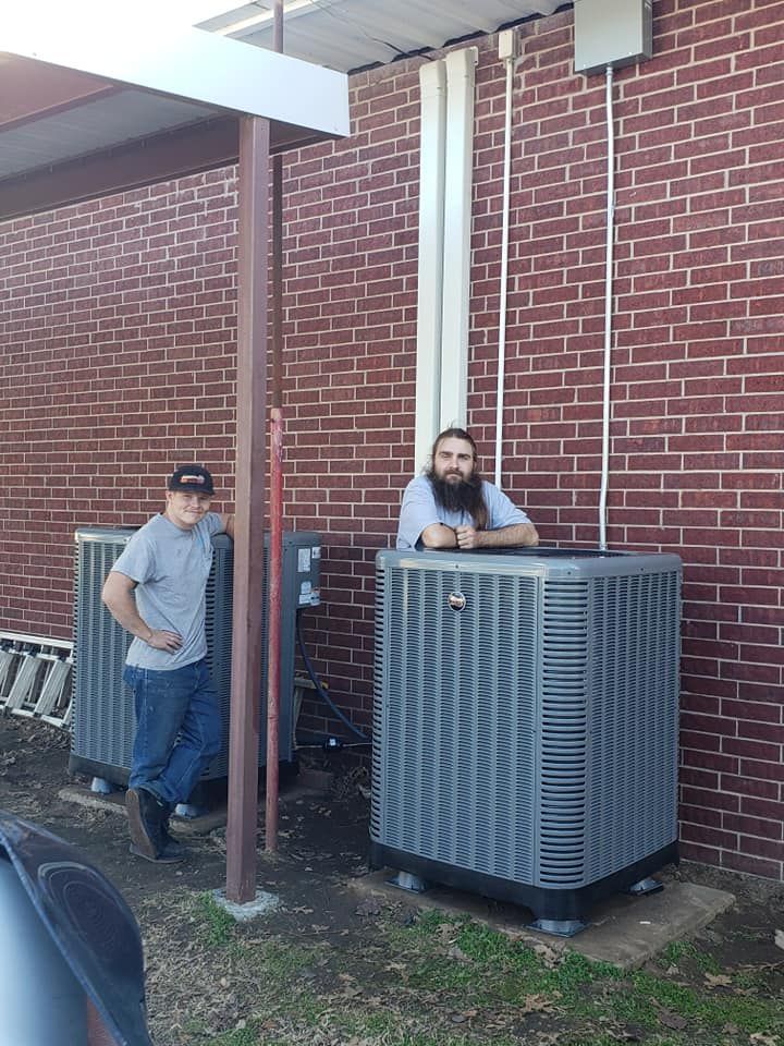 Two men are standing next to two air conditioners on the side of a brick building.