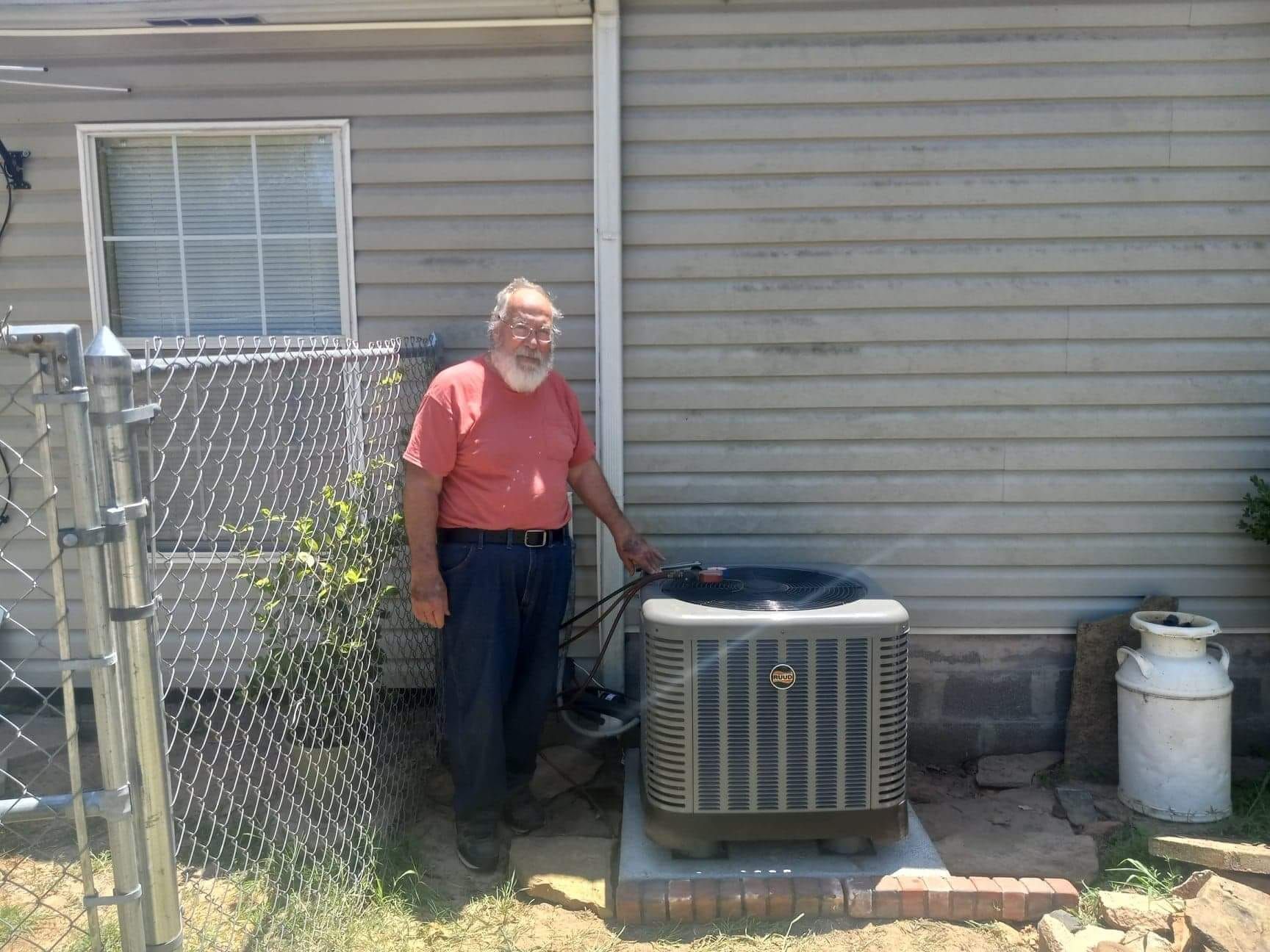 A man in a red shirt is standing next to an air conditioner outside of a house.