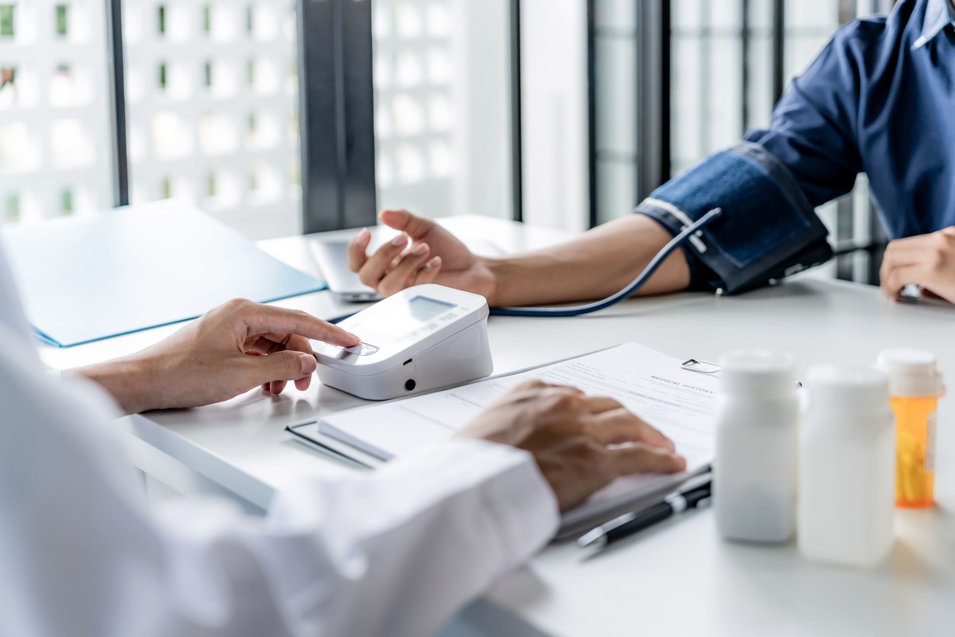 Doctor taking a patient's blood pressure. The patient sits at a desk with a cuff on their arm while the doctor operates the blood pressure monitor.