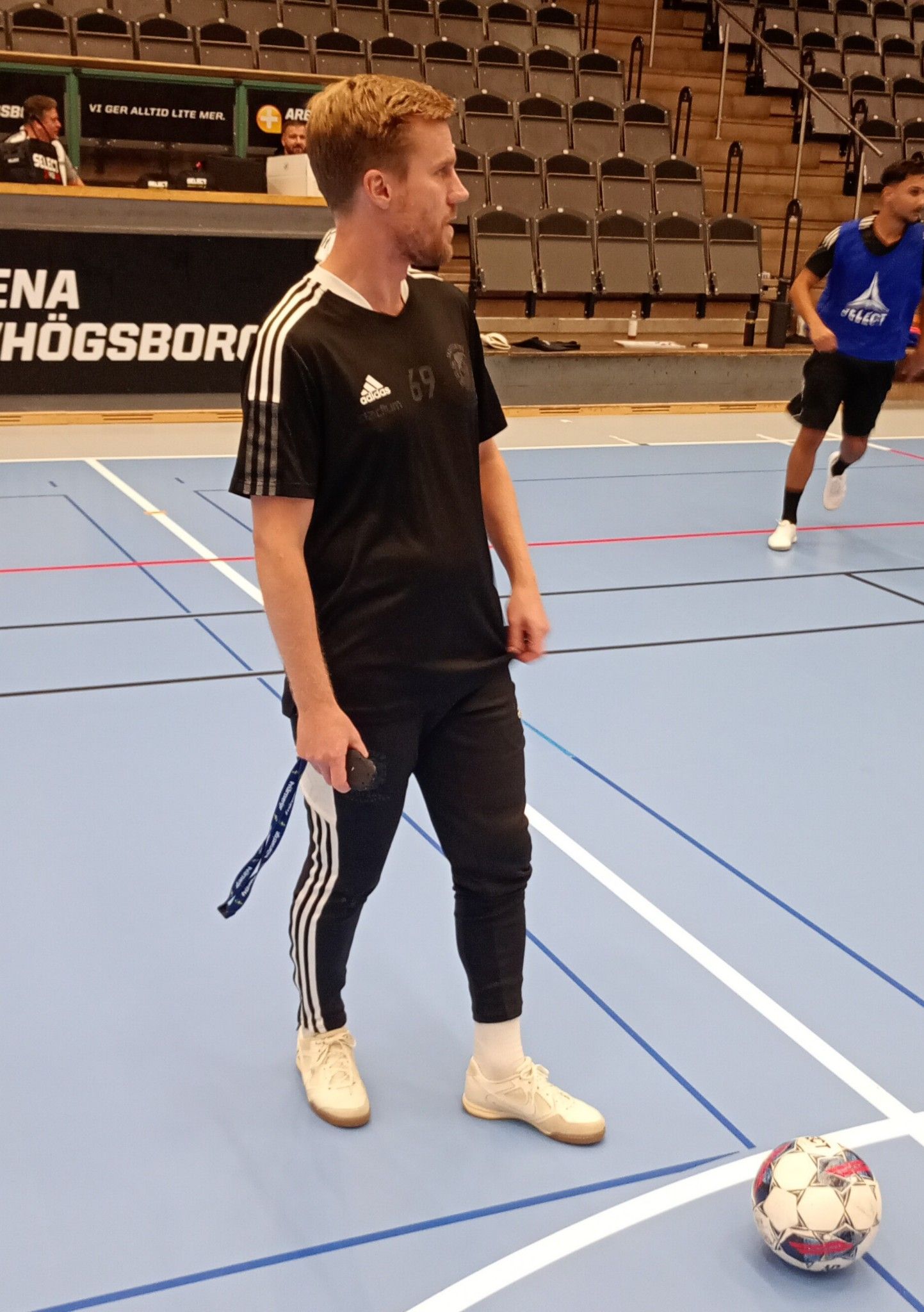 Man in black standing on court with a soccer ball, another player in the background.