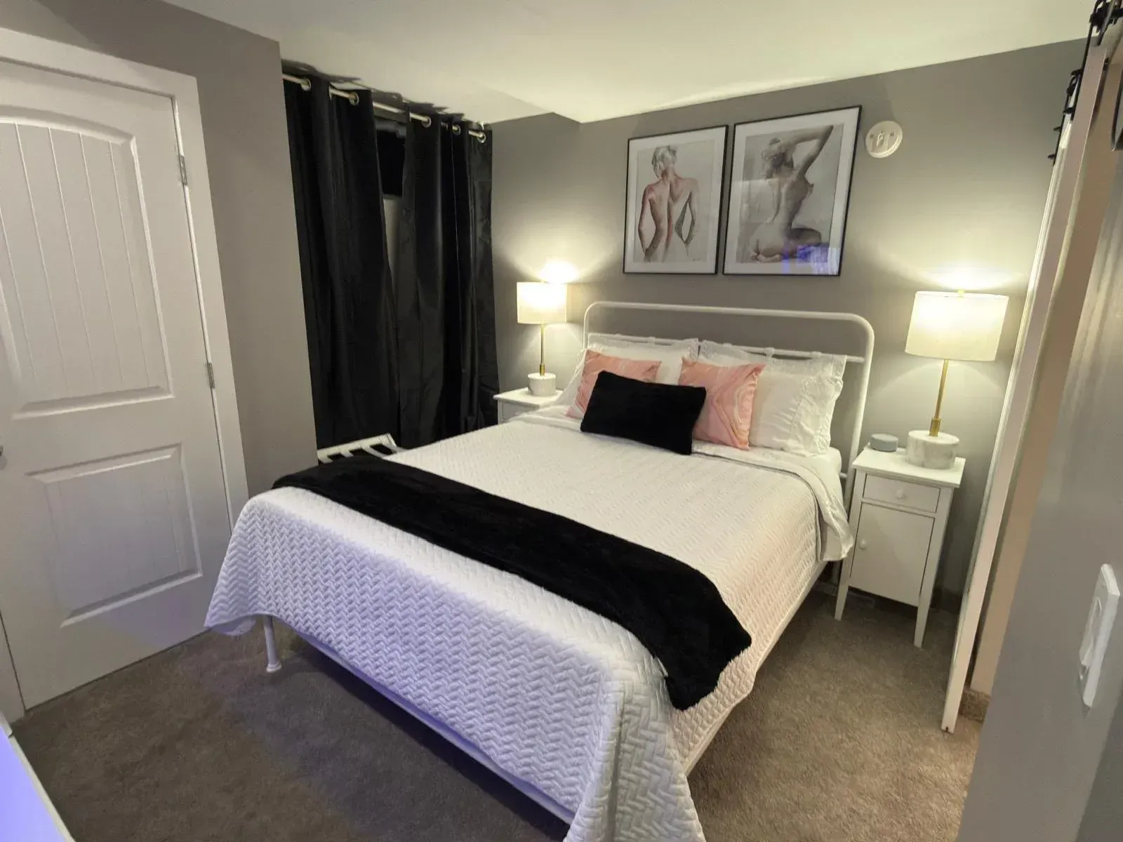 Bedroom with a neatly made bed, black curtains, bedside lamps, and framed artwork on gray walls.