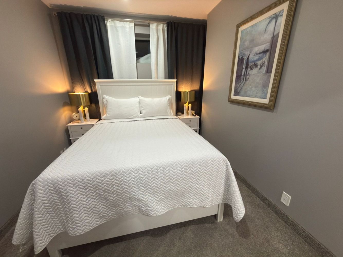 Small bedroom with a white bed, bedside lamps, dark curtains, and a framed painting on the wall.