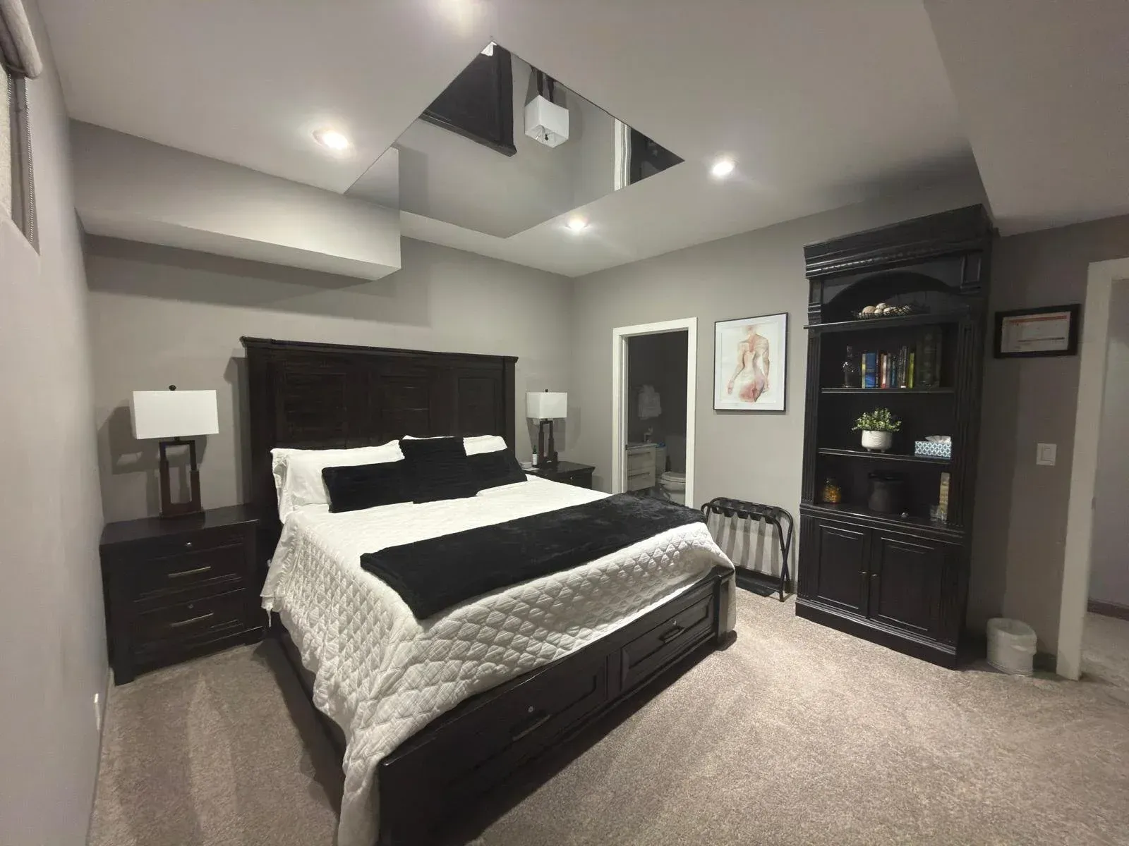 Modern bedroom with dark wood bed, mirrored ceiling panel, beige carpet, and black dresser and shelving unit