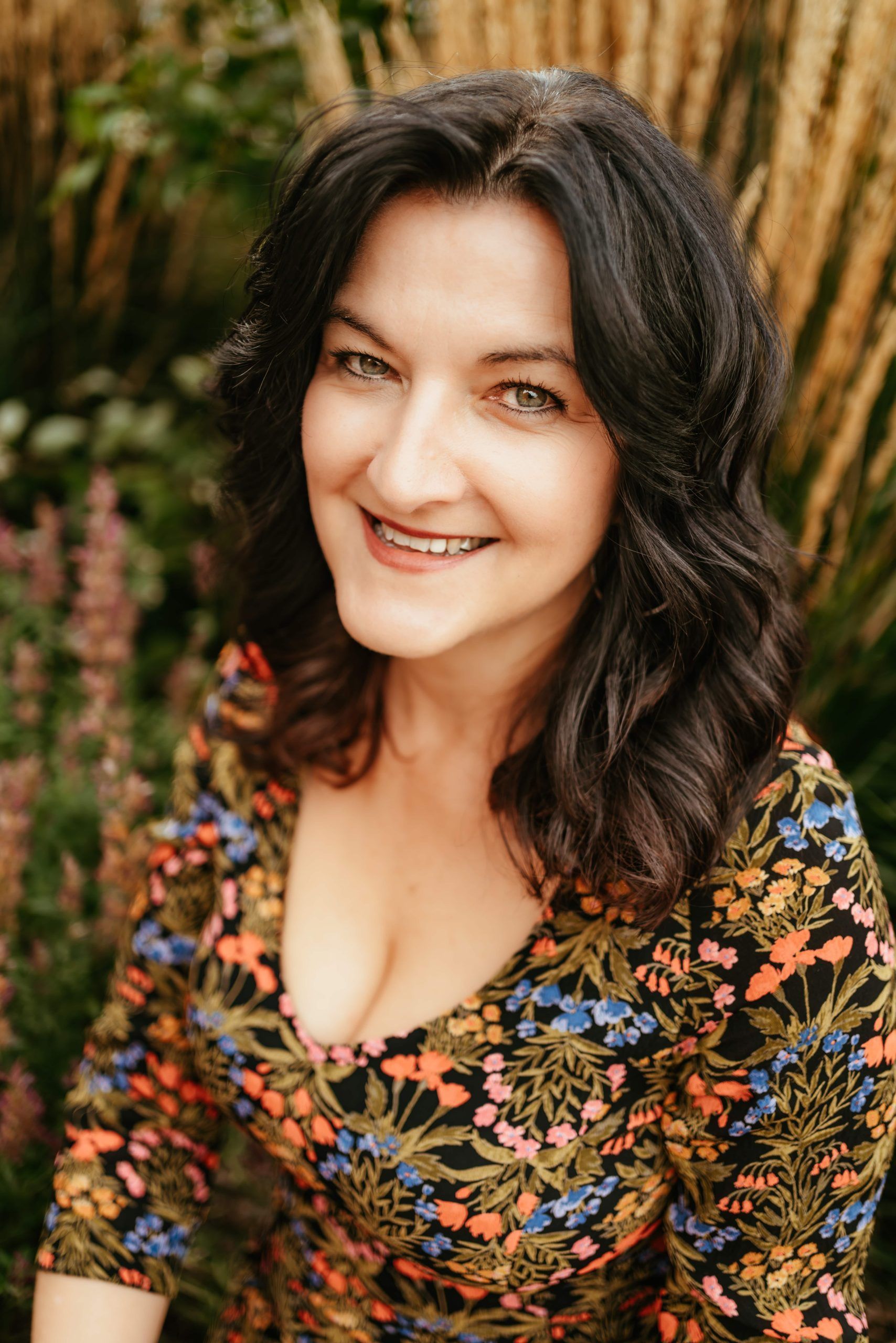 A smiling person with dark hair wears a floral print dress while standing outdoors among tall grasses.