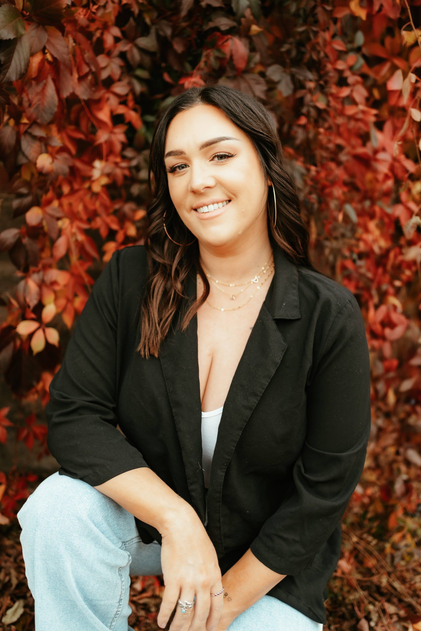 A smiling person wearing a black blazer and light blue jeans, posing in front of vibrant red autumn leaves.