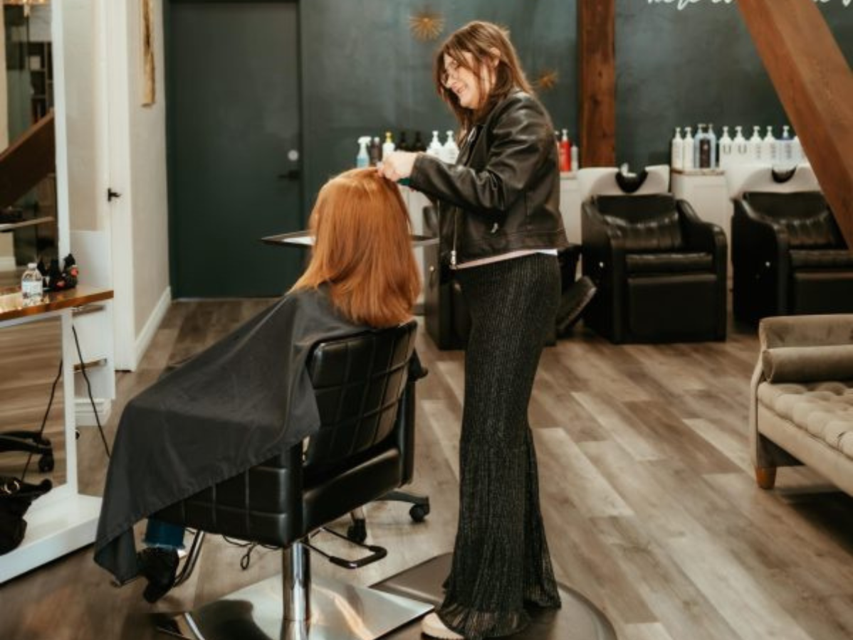 A hairstylist works on a client’s copper-colored hair in a salon with wooden floors and black leather styling chairs.