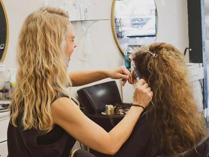 A hairstylist works on a client’s copper-colored hair in a salon with wooden floors and black leather styling chairs.