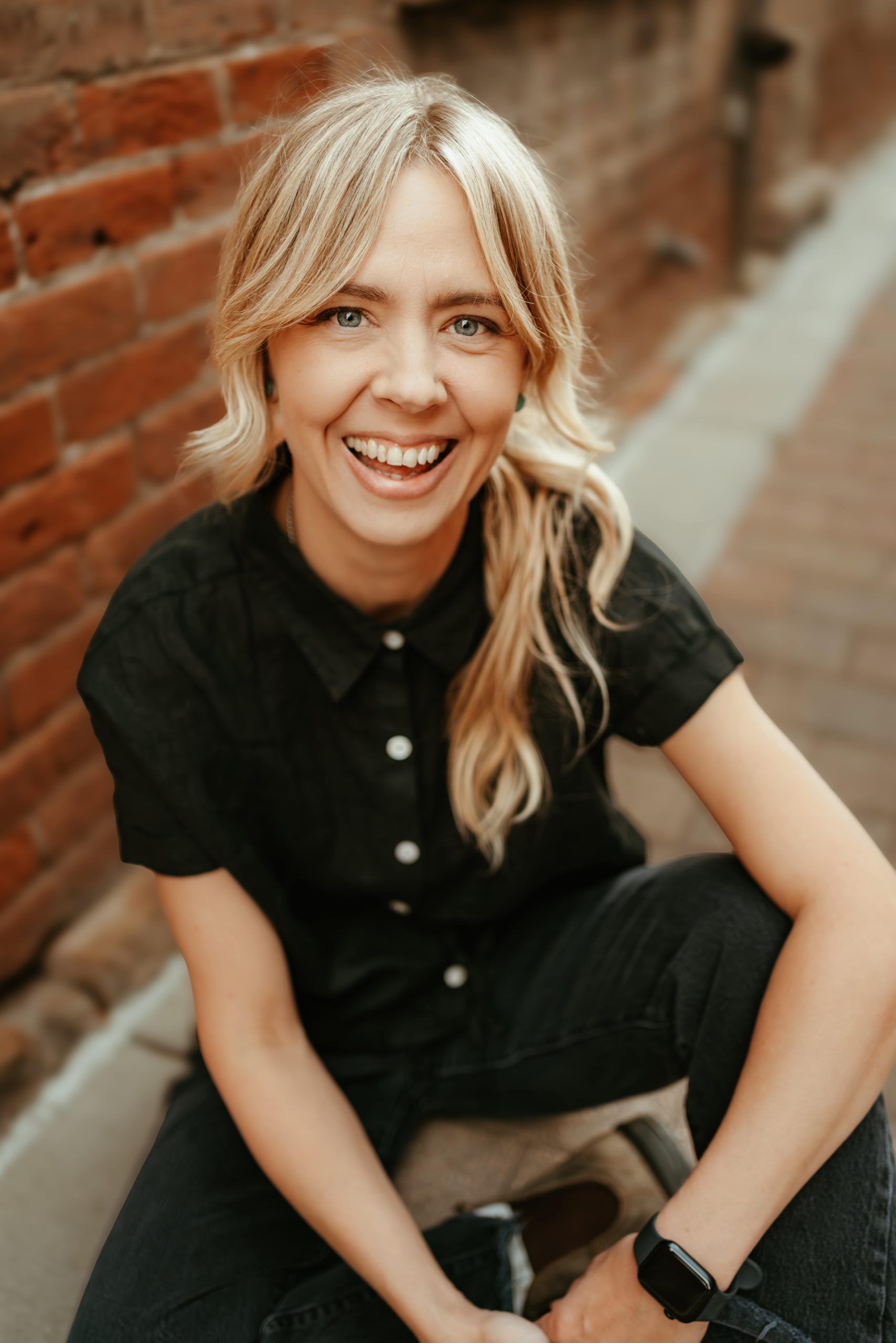 A person with blonde hair laughs while sitting against a brick wall, wearing a black shirt and a watch on their wrist.