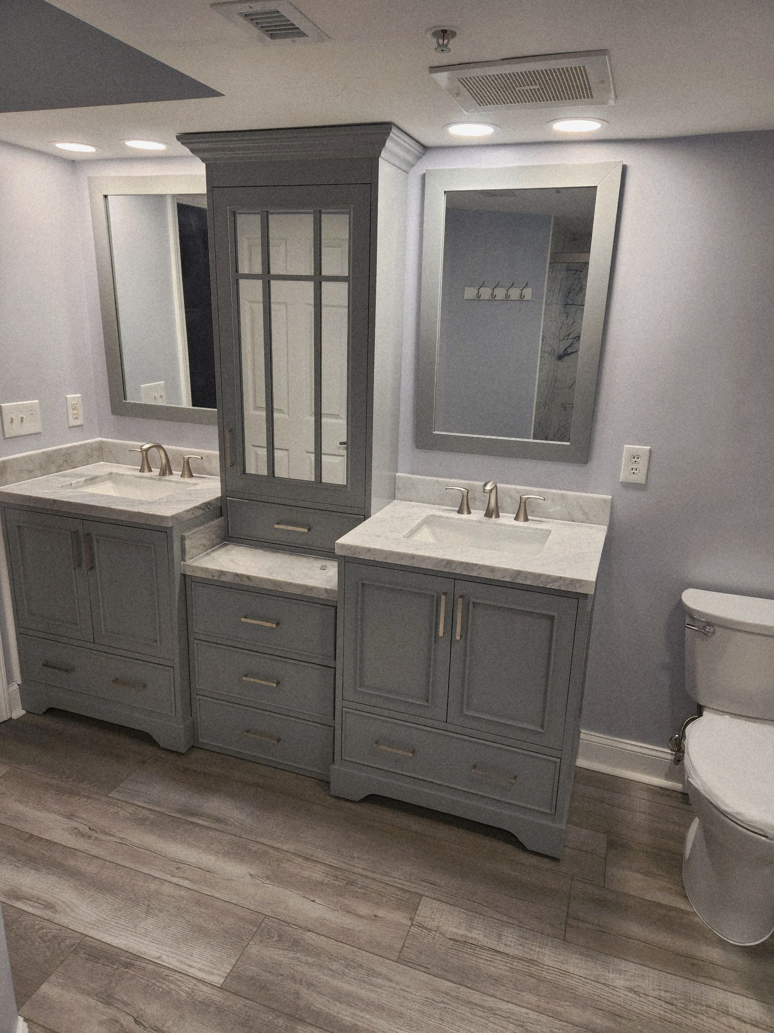 Bathroom vanity with two mirrors, gray cabinets, white sinks, and wood-look flooring under bright lighting