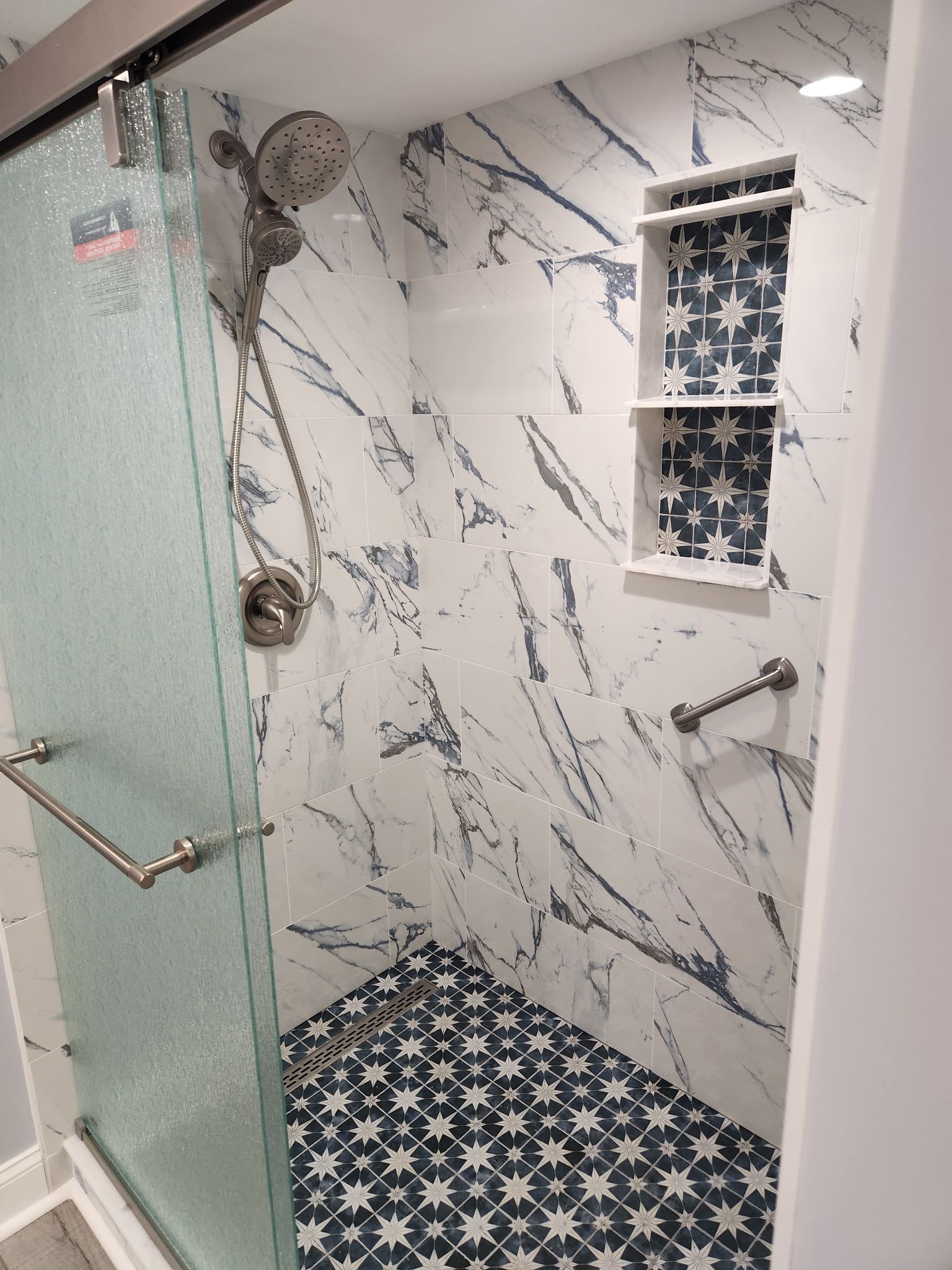 Small tiled shower with glass door, marble-look walls, black-and-white patterned floor, and a tiny window.