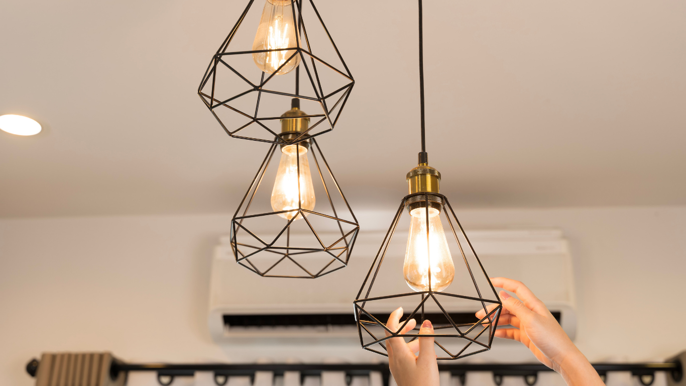 Three geometric pendant lights with warm bulbs hanging indoors, one being adjusted by a hand.