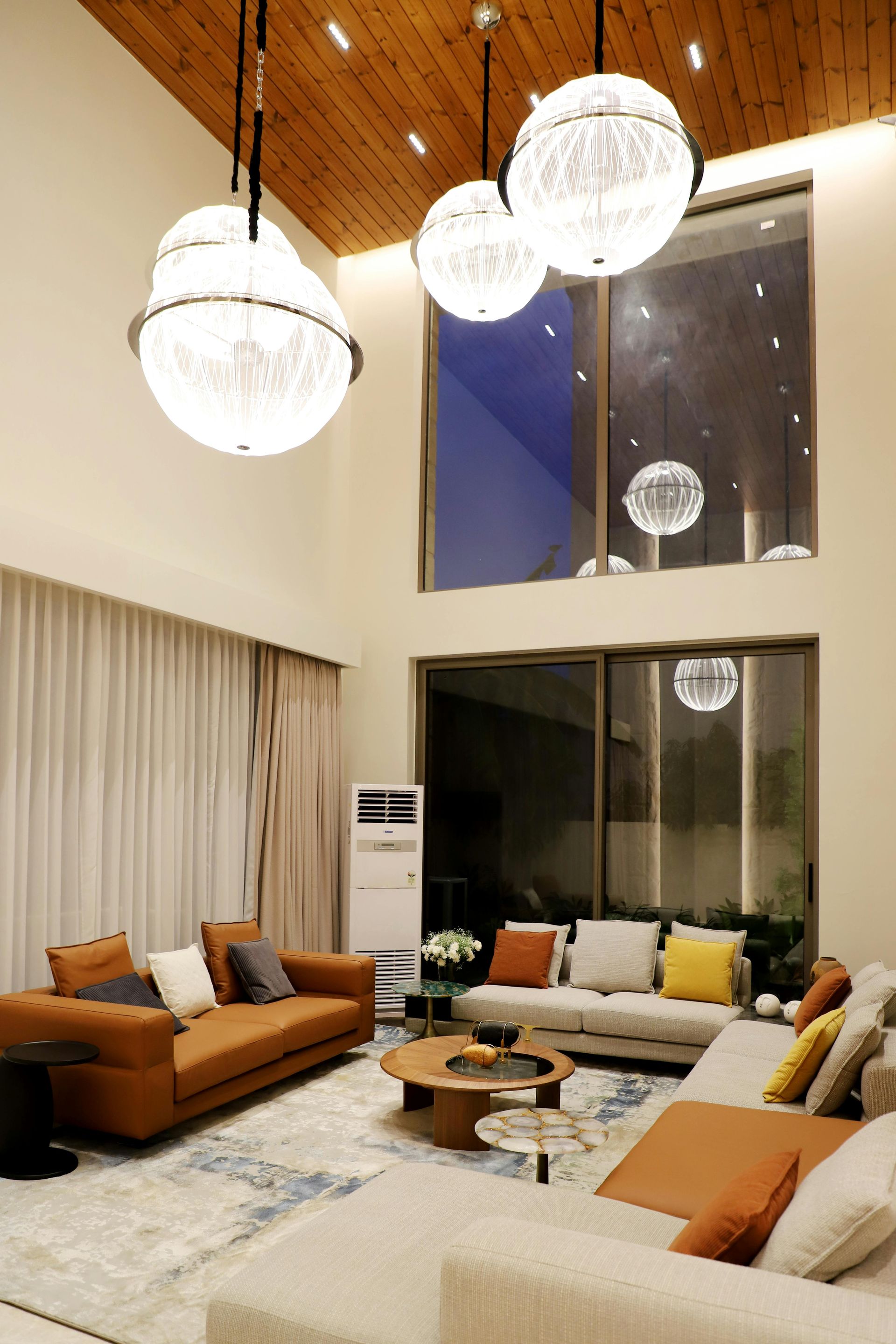 Bright modern living room with white sofas, tan chairs, round coffee table, and hanging globe lights under a wood ceiling