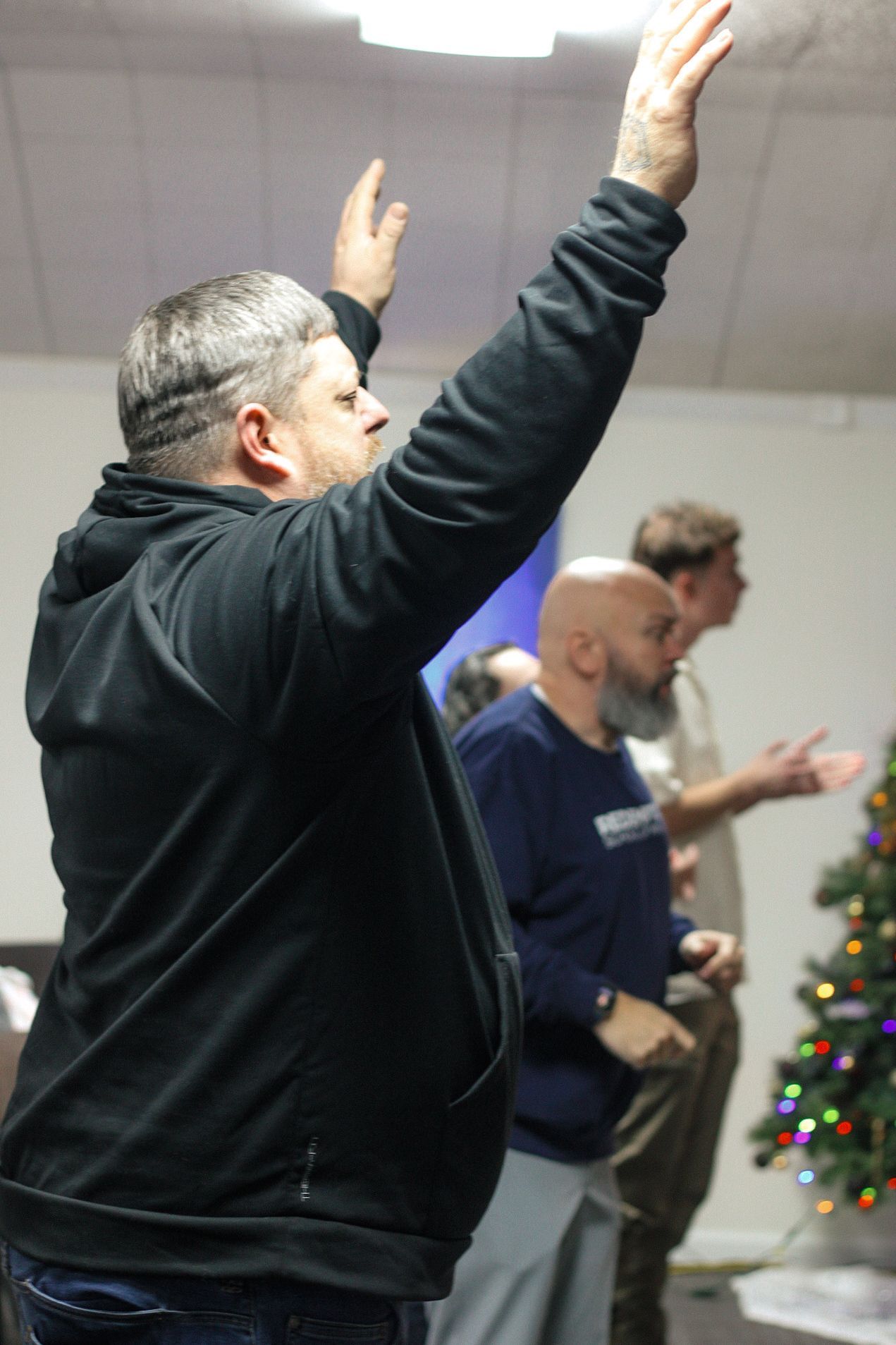 A man is raising his hands in the air in front of a christmas tree.
