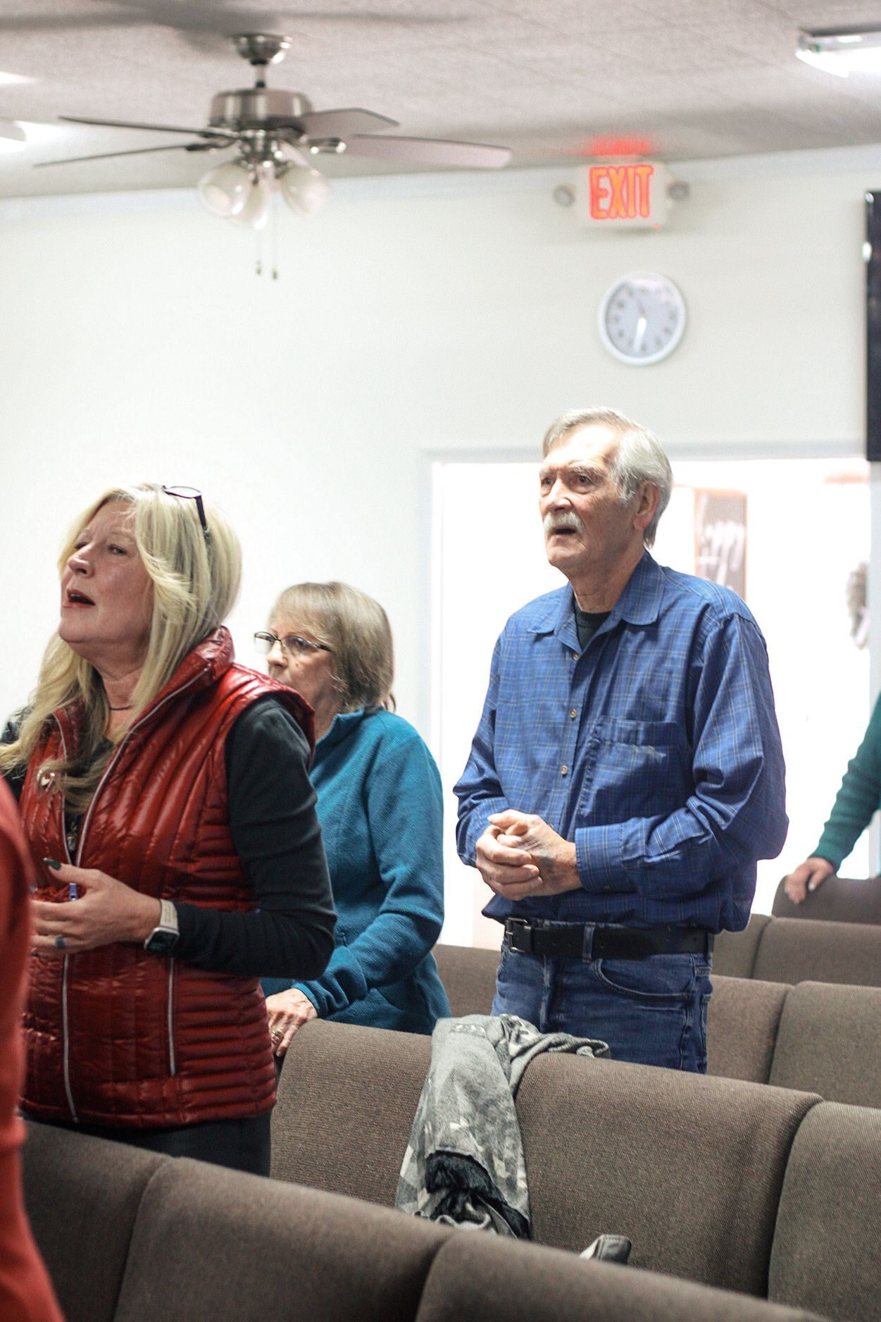 A group of people are sitting in a room with a red exit sign on the wall