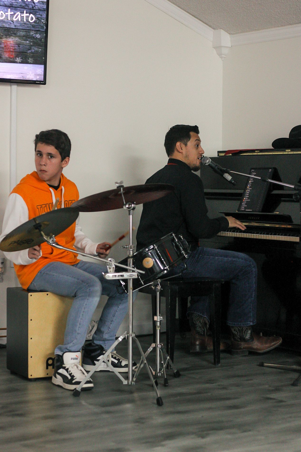 Two young men are playing drums and a piano in a room.