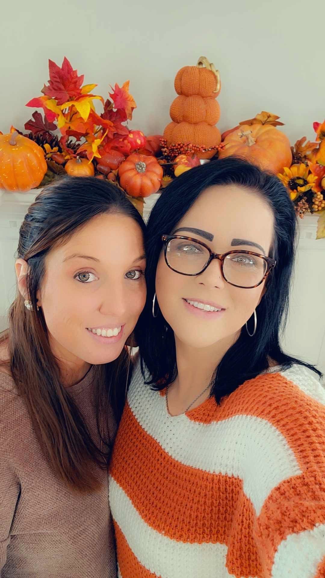 Two women are posing for a picture in front of pumpkins and leaves.