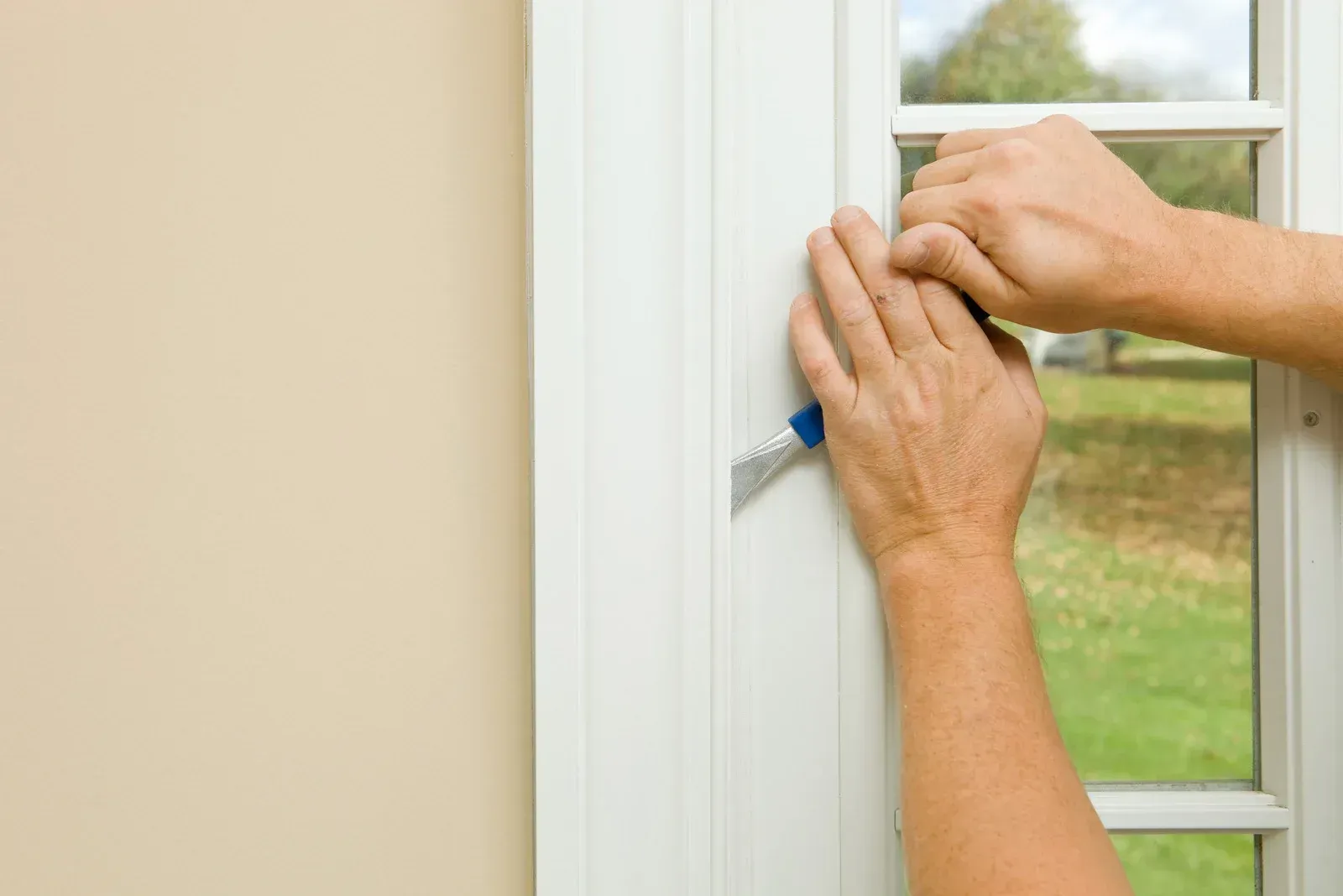 Hands using a utility knife to scrape paint from a white window frame, outdoor background.