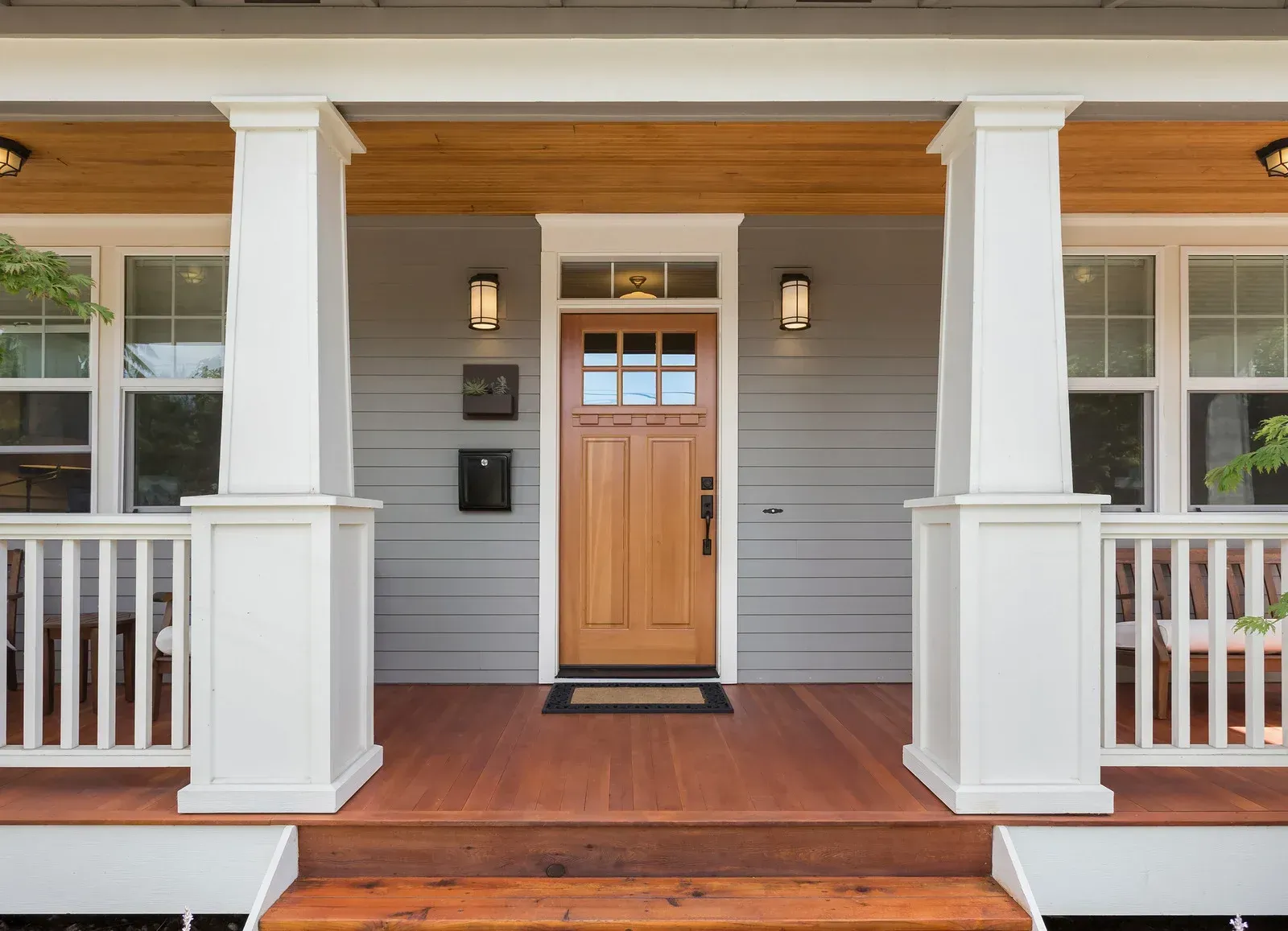 Front porch of a gray house with white columns, a wooden door, and a brown wood floor.