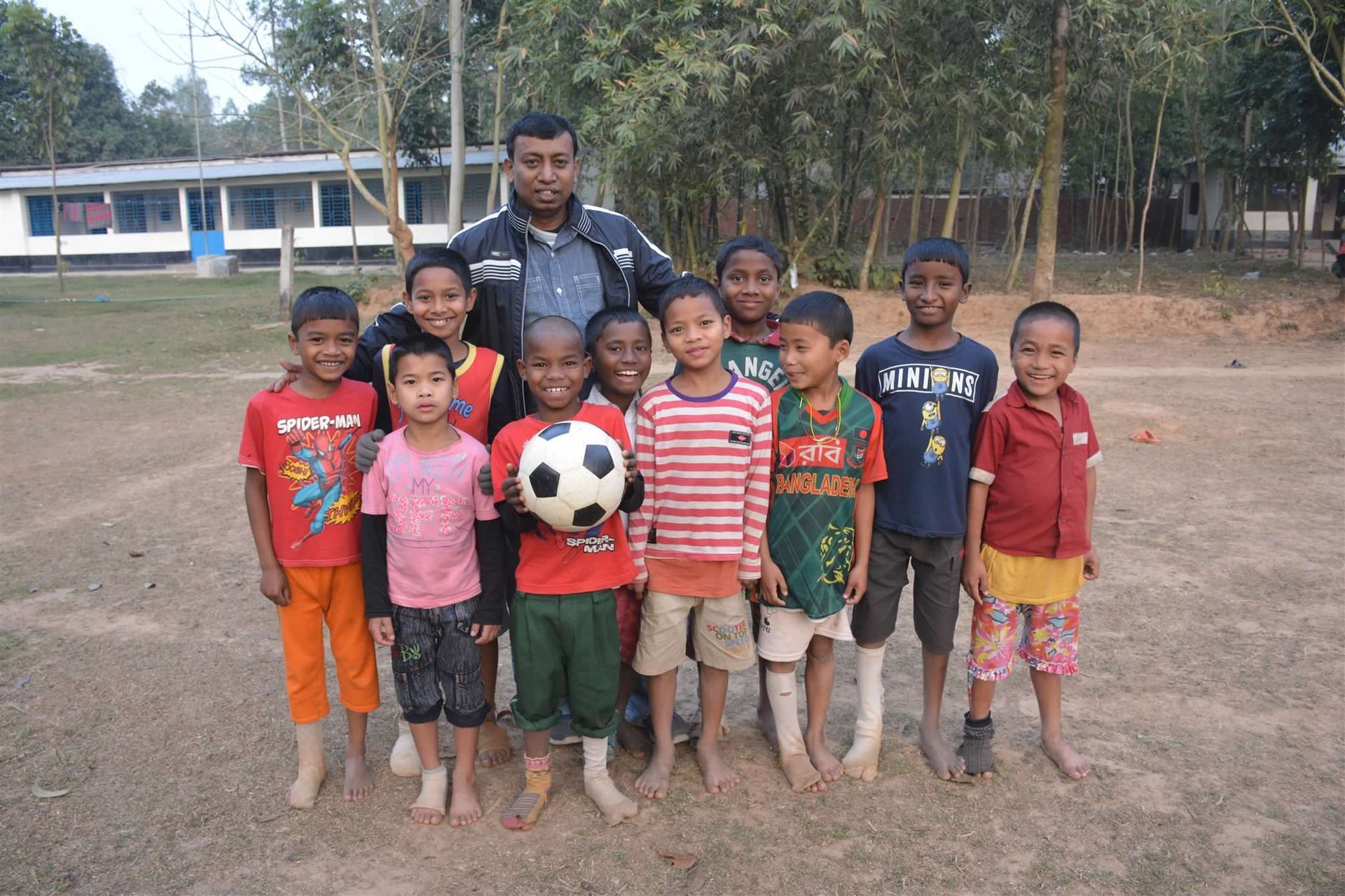 A group of children and an adult standing outdoors on a dirt field, with one child holding a soccer ball.