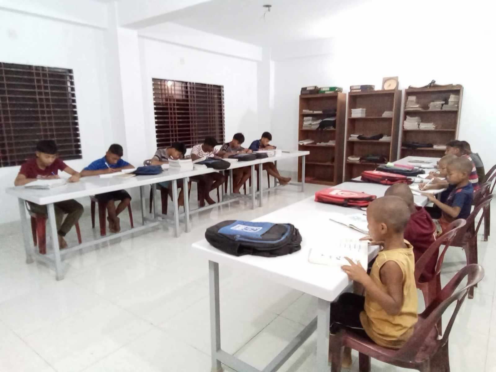 Students sit at desks in a bright, white classroom, studying at tables with books and backpacks.