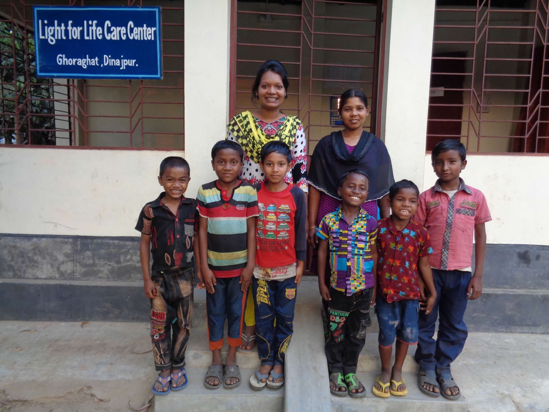 Two caregivers stand behind six smiling children in front of a building sign for the Light for Life Care Center.