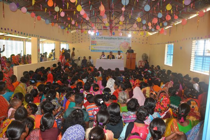 A large group of people gathers in a decorated hall for a formal event with speakers at a table on a stage.