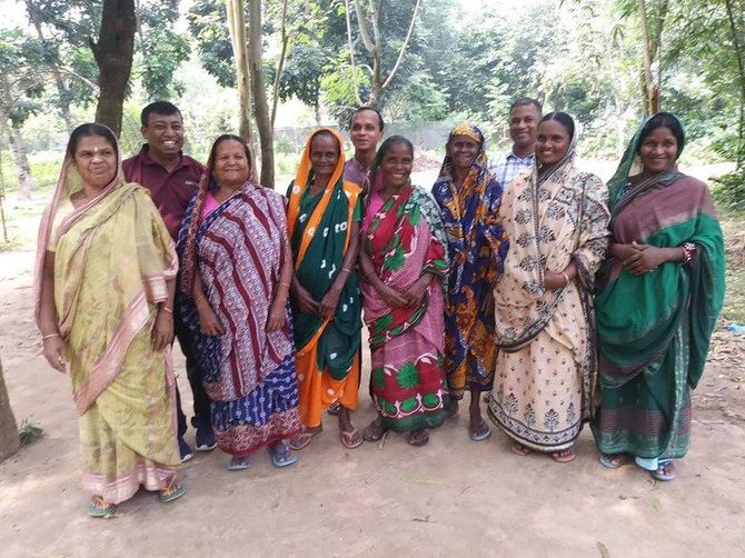 A group of ten people stand outdoors in a wooded area, posing together and smiling for the camera.
