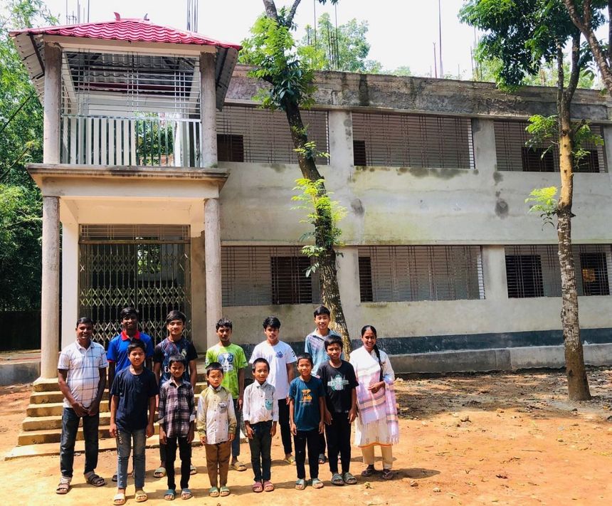 A group of people standing in front of a two-story building with a covered entrance in a rural setting.