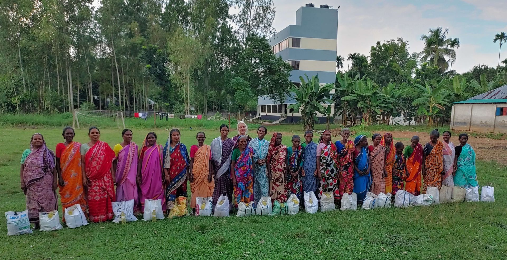 A group of people standing in a grassy field, each holding a white bag, with trees and a building in the background.