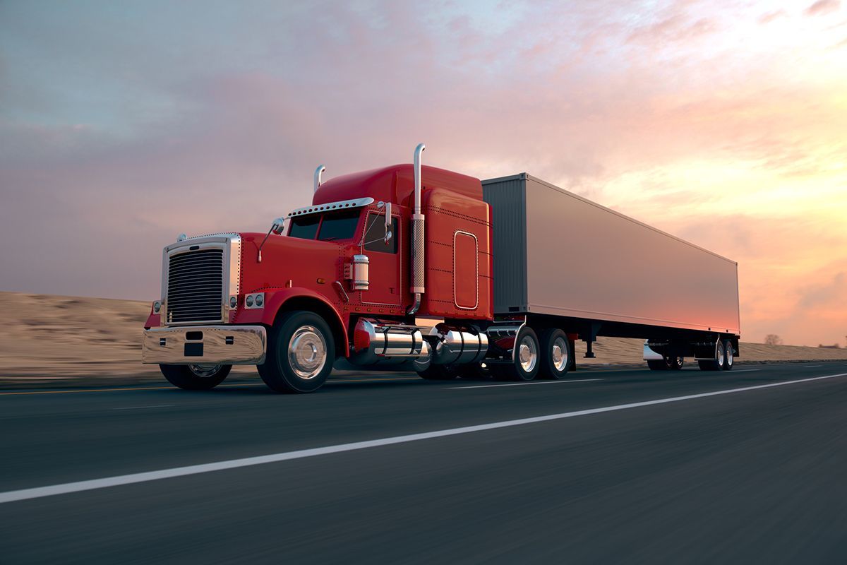 Red semi-truck with trailer driving on a highway at sunset.