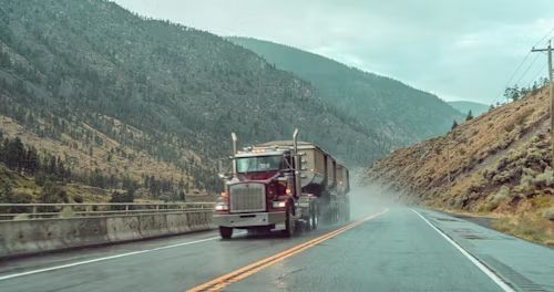 Red semi-truck driving on a wet mountain highway with mist and hills in the background