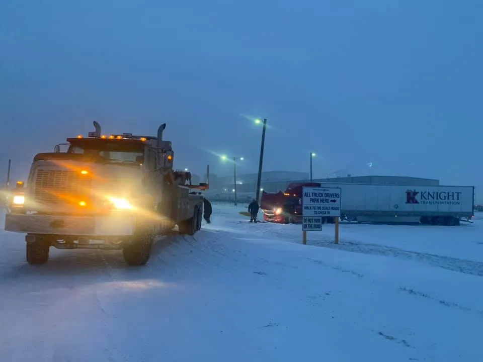 Tow truck on snowy roadside at dusk with lights on, near a KNIGHT trailer and streetlights
