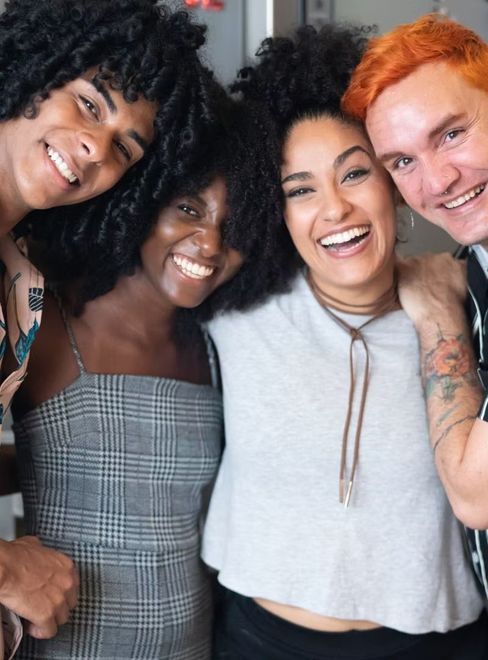 Four friends smiling closely together in casual clothes, posing for a selfie indoors.