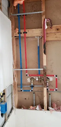 Piping and plumbing fixtures installed inside a bathroom wall. Blue and red pipes connected to a faucet.