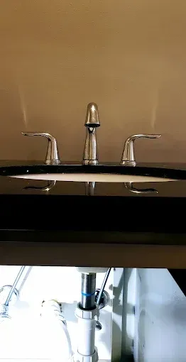 Bathroom sink with chrome faucet and handles, reflected in a dark counter, with plumbing visible below.