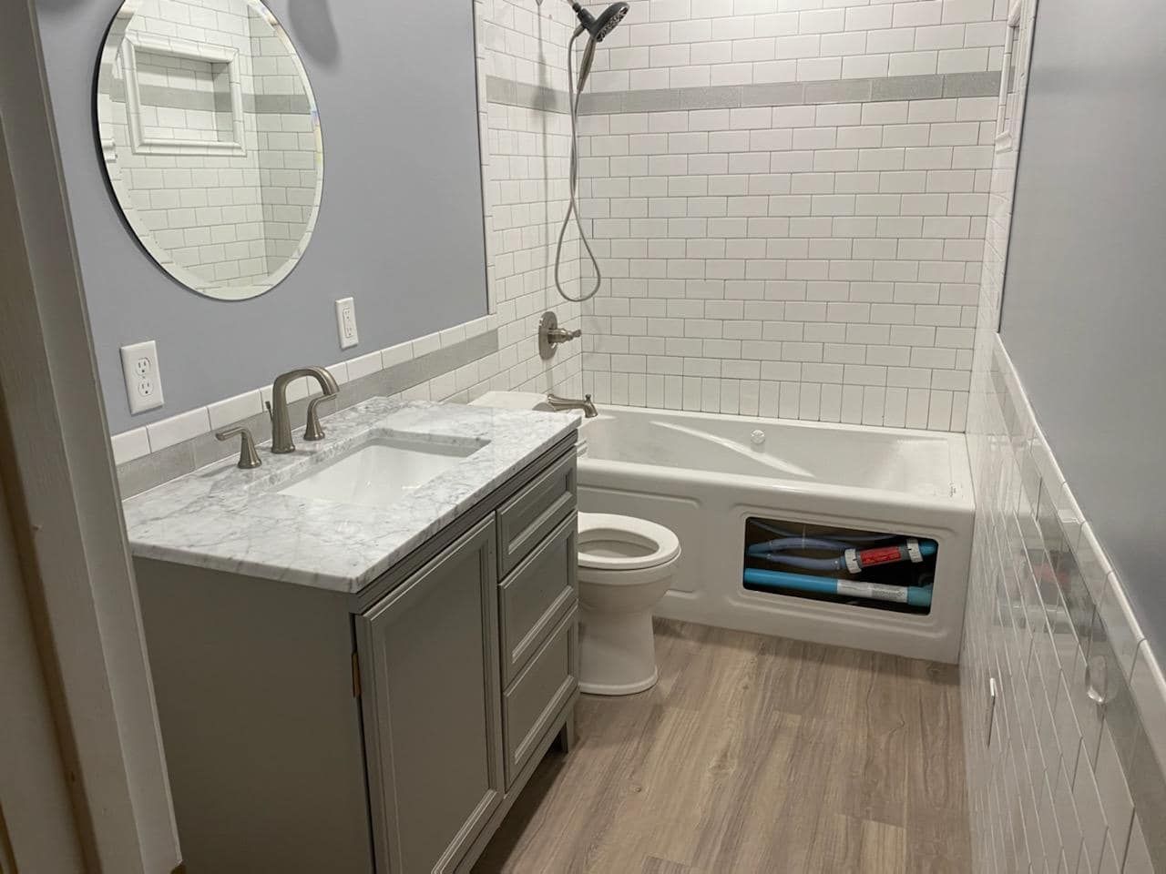 Bathroom with white subway tile shower, grey vanity, round mirror, and wood-look floor.