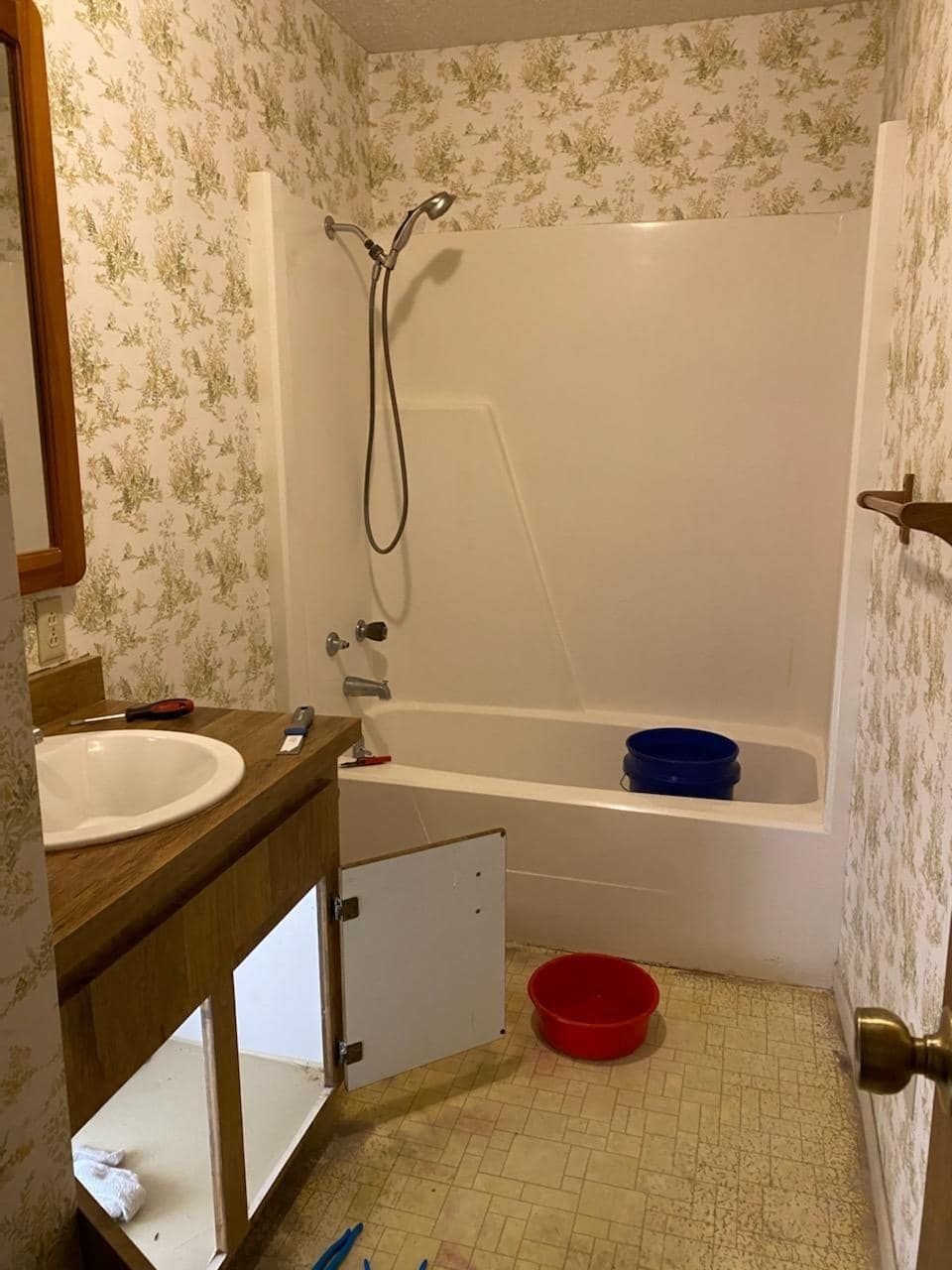 Bathroom with floral wallpaper, a white tub/shower, a vanity with an open cabinet, and a red and blue bucket on the floor.