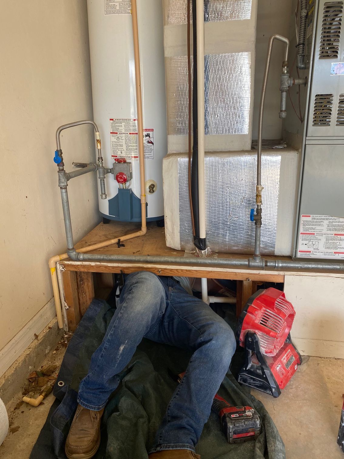 A person in blue jeans is working near a furnace and water heater. Red tool on the floor.