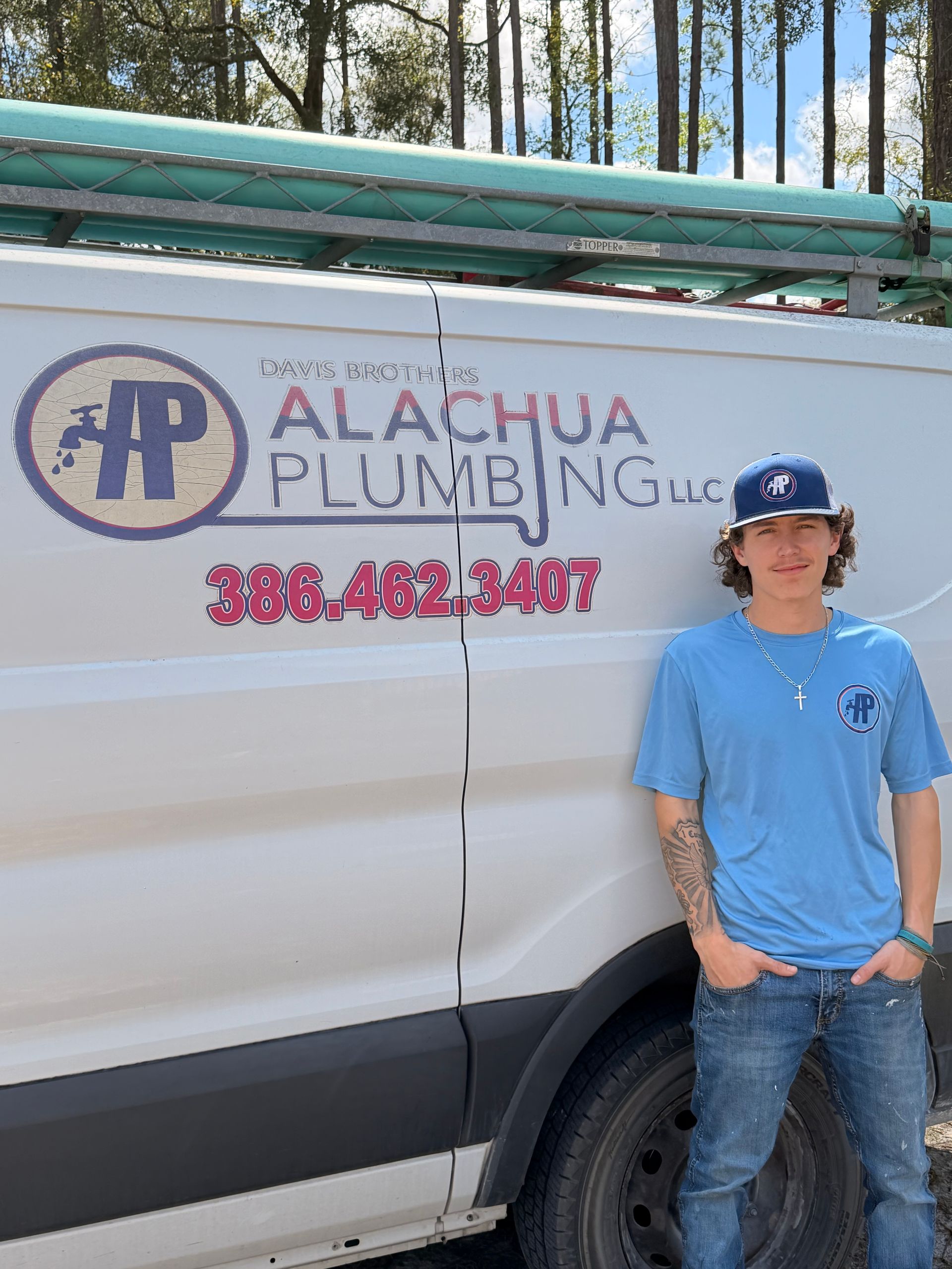 Man in a baseball cap and long-sleeved shirt, posing next to a plumbing truck with logo and phone number.