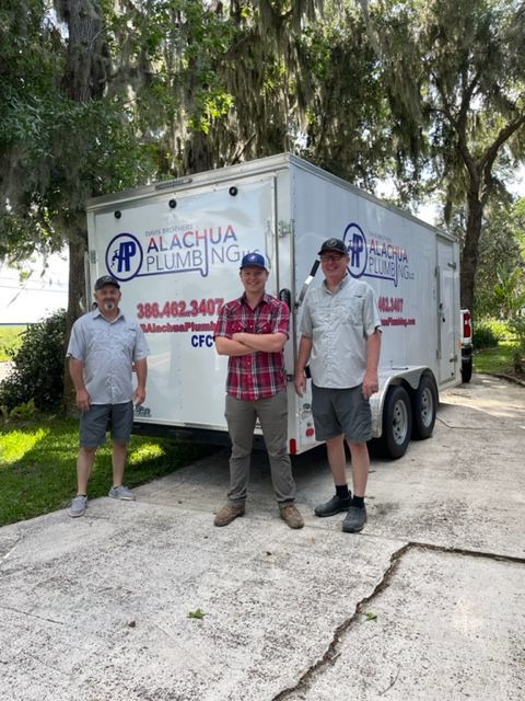 Three men stand in front of a white trailer for Alachua Plumbing. The trailer is parked on a driveway.