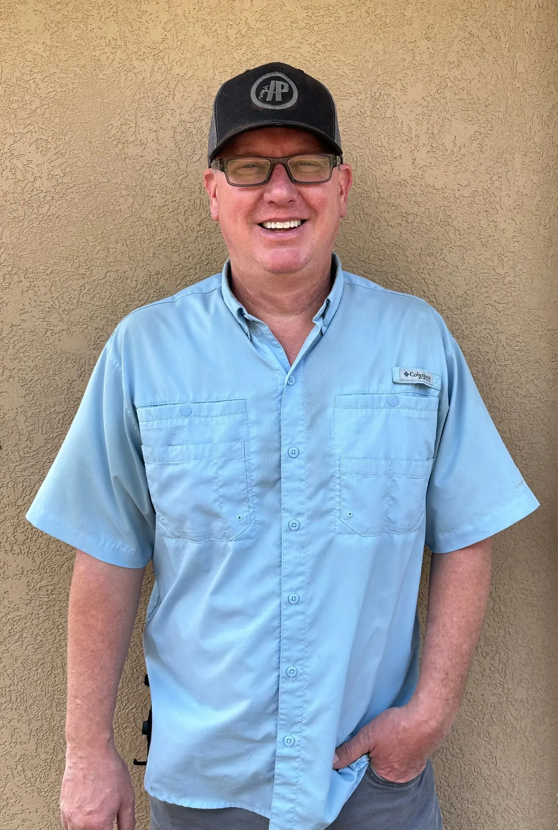 Man in blue shirt, glasses, and hat smiles, leaning against a tan wall, hand in pocket.