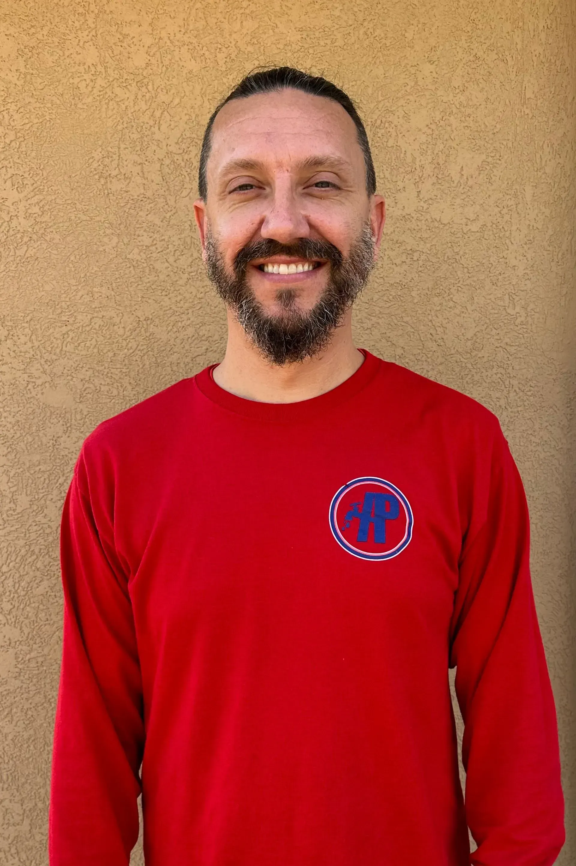 Man with beard in red shirt smiles at camera, standing outside.