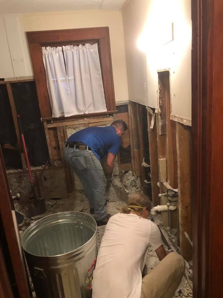 Two people are demolishing a bathroom. One uses a tool, the other works on plumbing. Debris covers the floor.