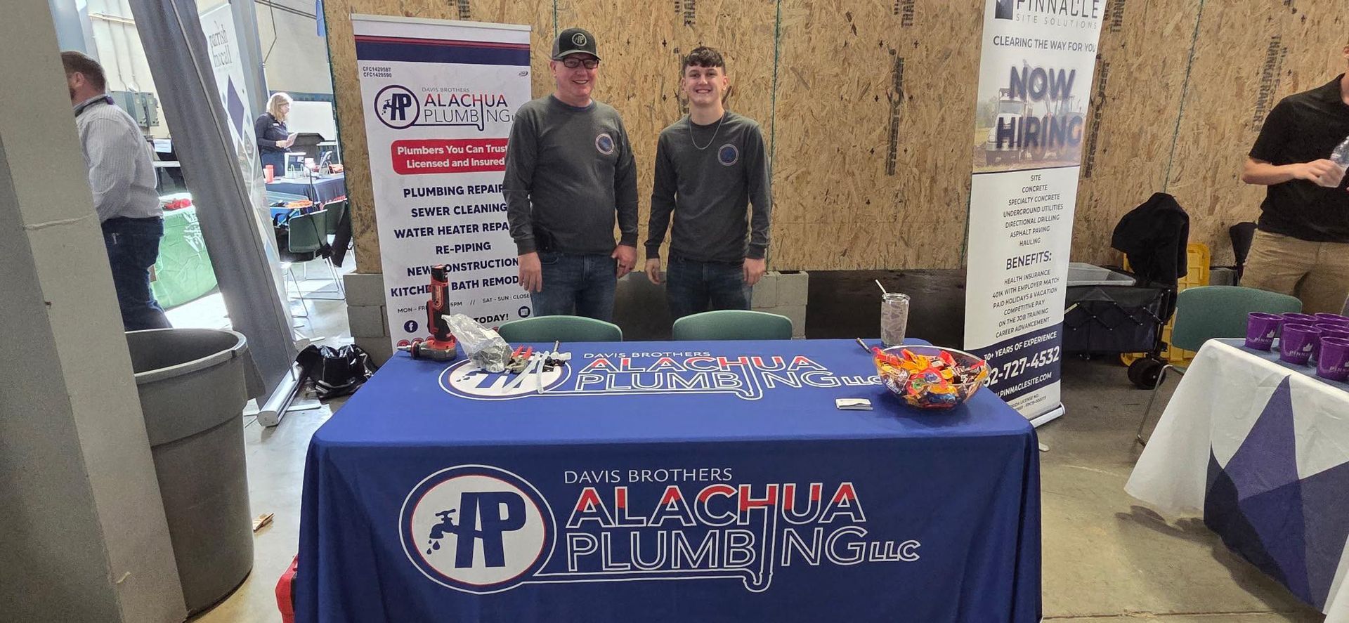 Two people stand behind a table with a company logo. They are at an event with banners.