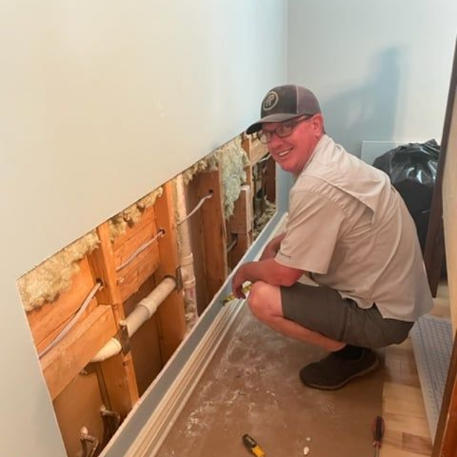 Man crouches by a hole in a wall, likely repairing plumbing. Exposed wood, wires, and insulation visible. Tools on the floor.