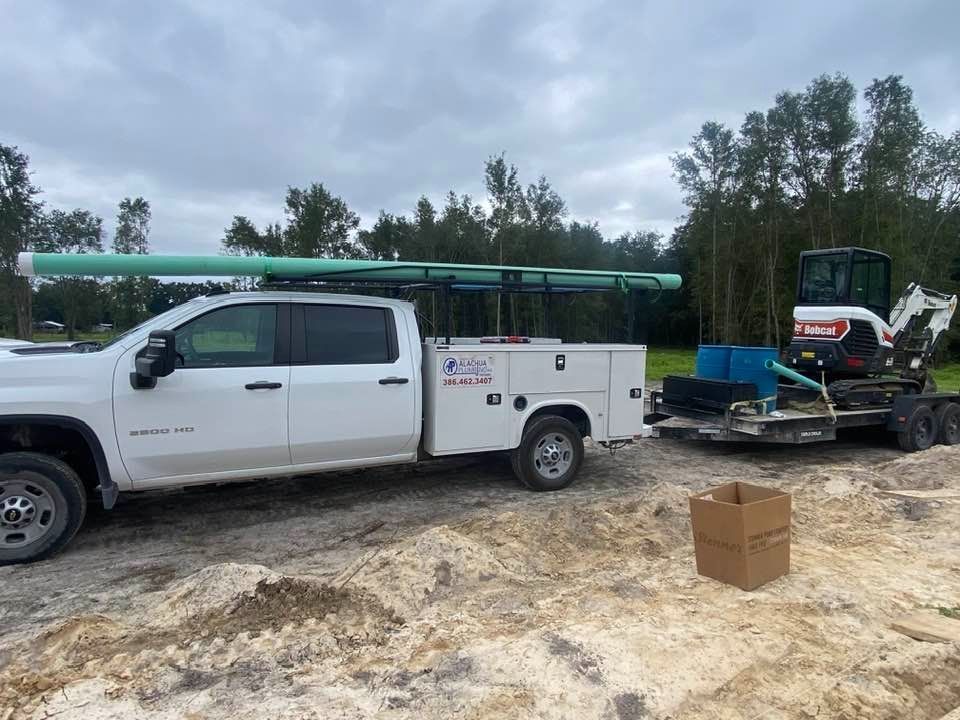 White work truck with pipes on the roof, a Bobcat excavator on a trailer, and a cardboard box on dirt.