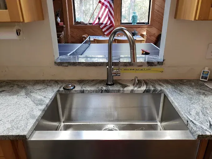 Stainless steel kitchen sink with faucet, granite countertop, and view into a room with a ping pong table.