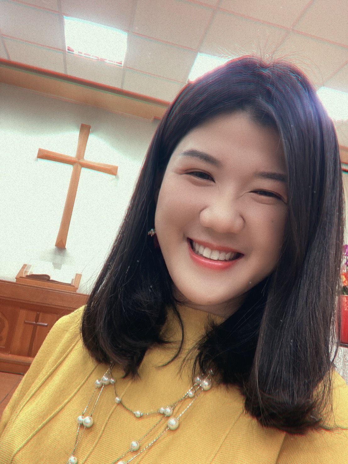 一位身穿黄色上衣、佩戴珍珠项链的微笑女子在房间里自拍，房间里的墙上挂着一个木制十字架。