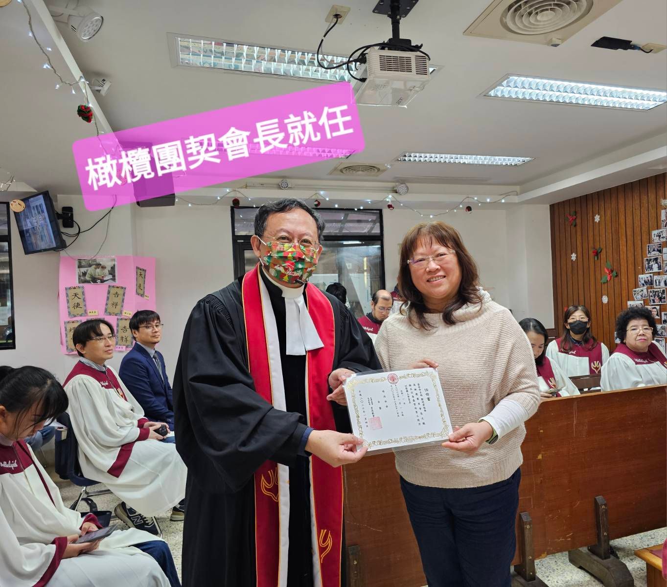 一位身穿神职人员长袍的人在一个房间里向一位女士颁发证书，房间里还有一群人，证书上覆盖着粉色文字。