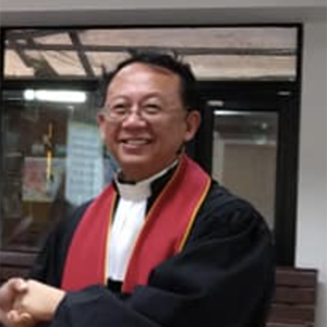 一位面带微笑的神职人员身穿黑色长袍，披着红色圣带，戴着白色神职领，在握手时做着手势。